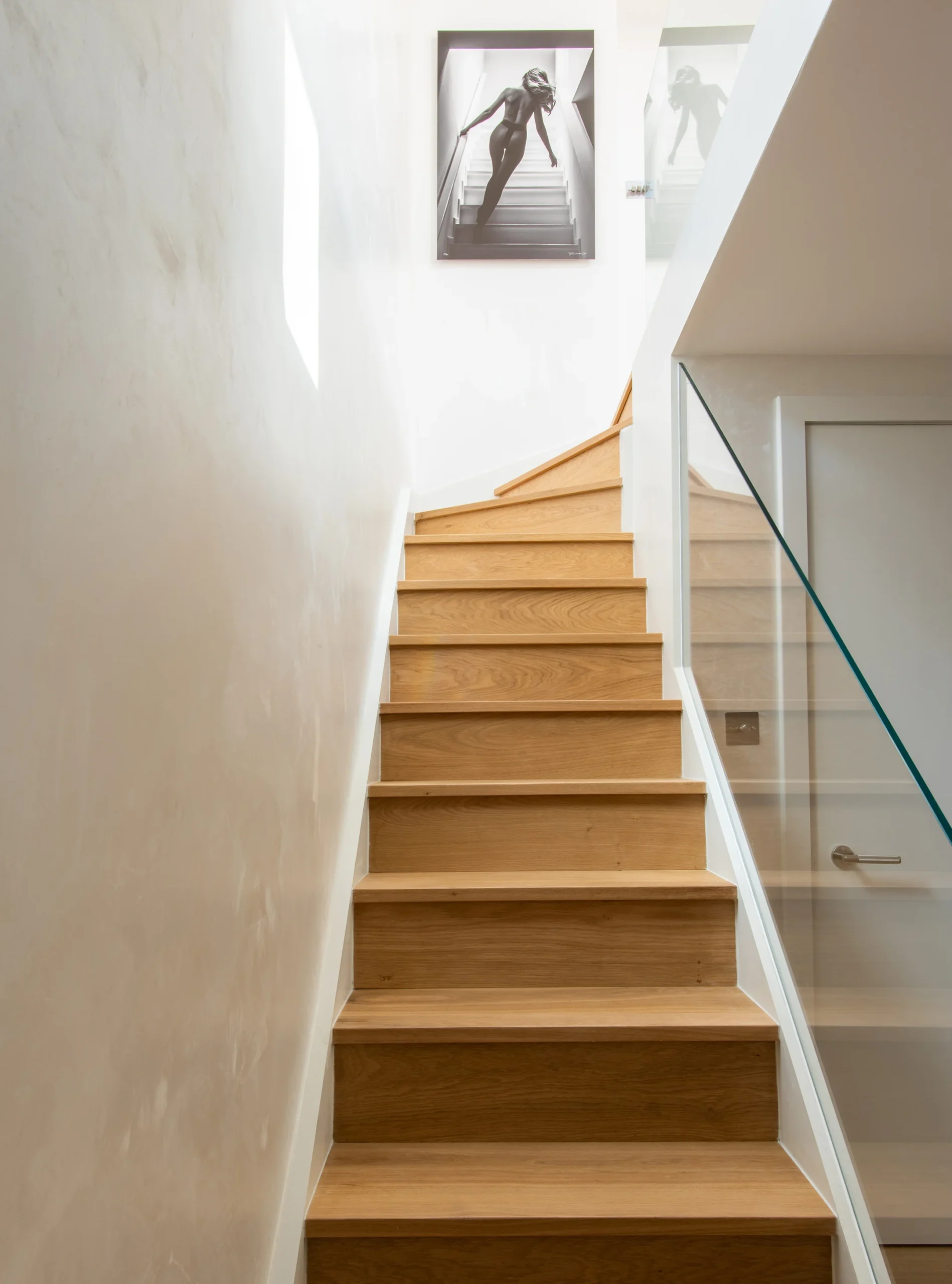 Oak staircase with frameless glass balustrade rising between white walls