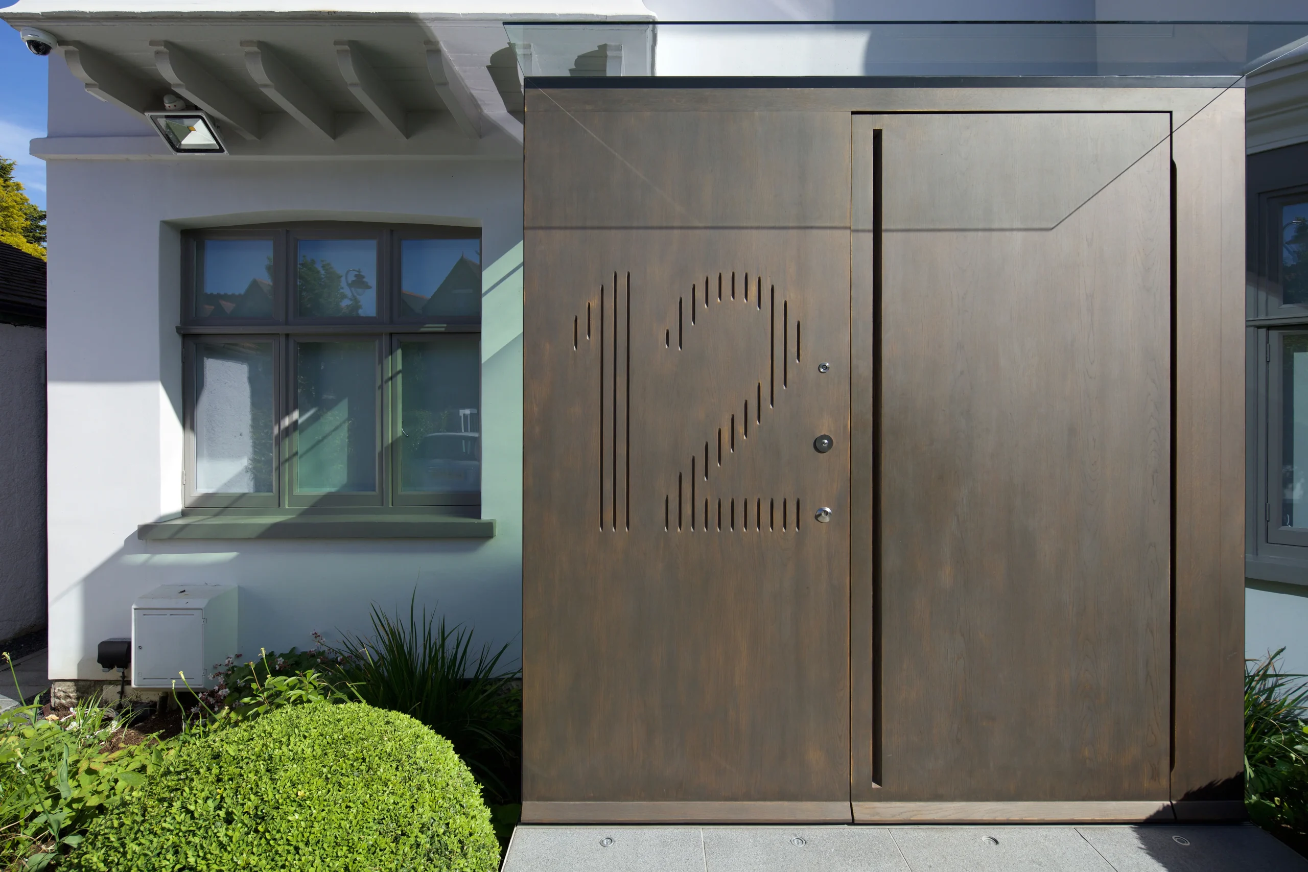 Bronze-clad front door with cut-out house number beside the rendered facade
