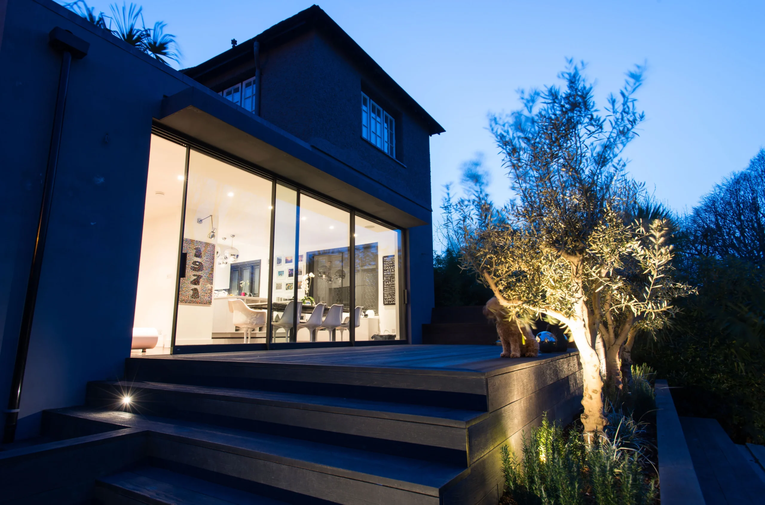Rear extension of a Willesden Green house at dusk, with full-height glazing, lit timber deck and olive tree